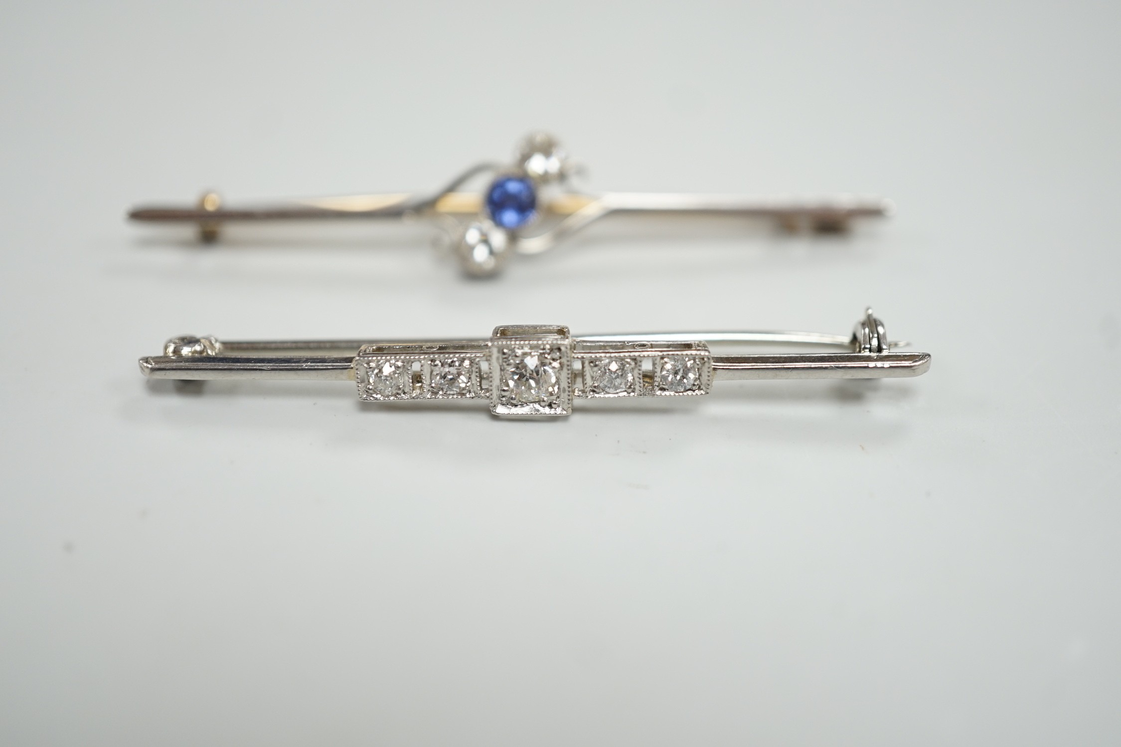 An early to mid 20th century white metal and five stone diamond set bar brooch, 50mm and a similar yellow metal, two stone diamond and single stone sapphire set crossover bar brooch, gross weight 7.1 grams.
